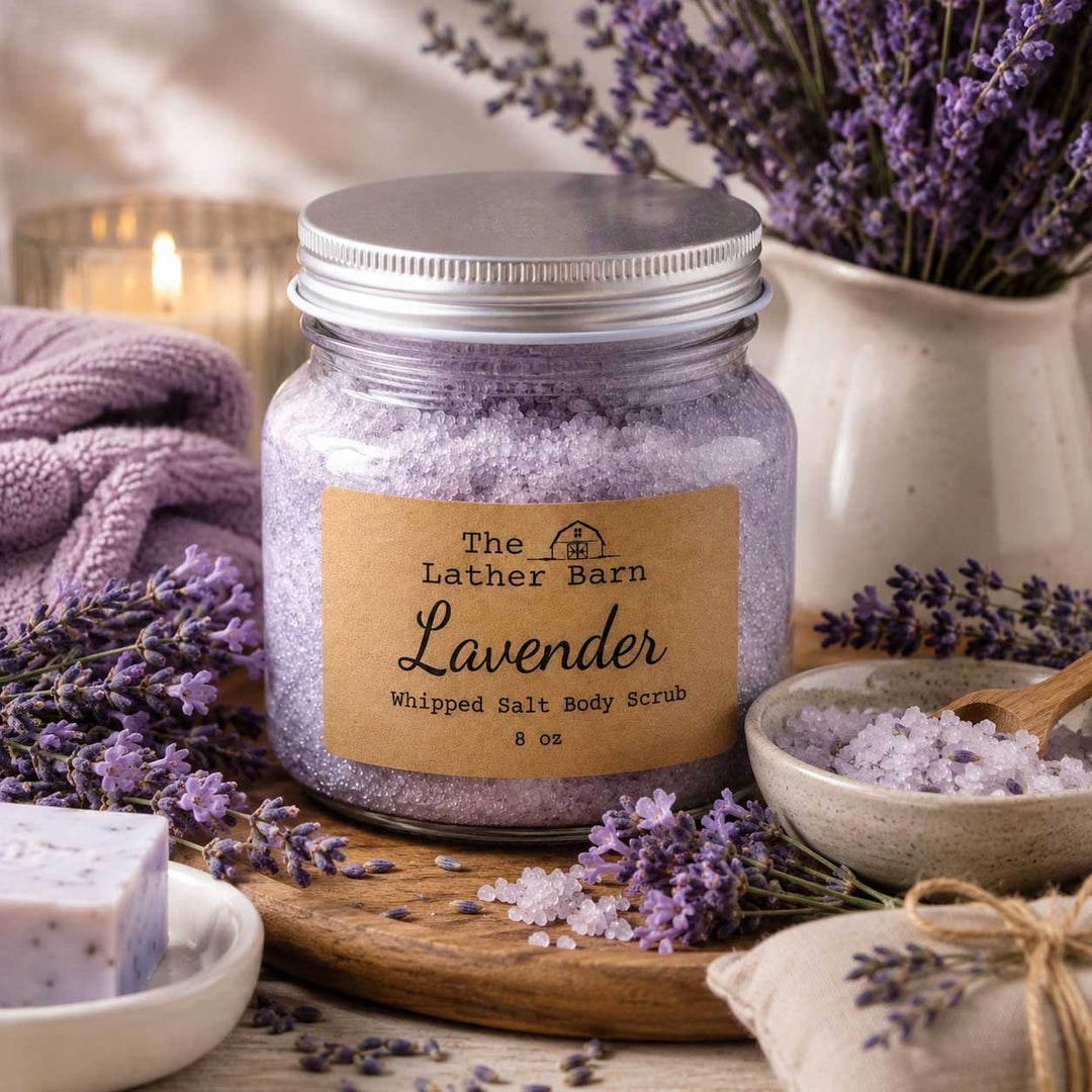A jar of lavender whipped salt body scrub by The Lather Barn sits on a wooden board surrounded by lavender sprigs, bowls of scrub, a folded towel, and a candle in a softly styled setting.