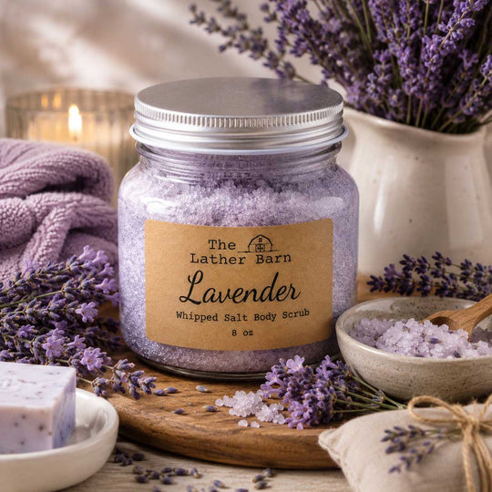 A jar of lavender whipped salt body scrub by The Lather Barn sits on a wooden board surrounded by lavender sprigs, bowls of scrub, a folded towel, and a candle in a softly styled setting.