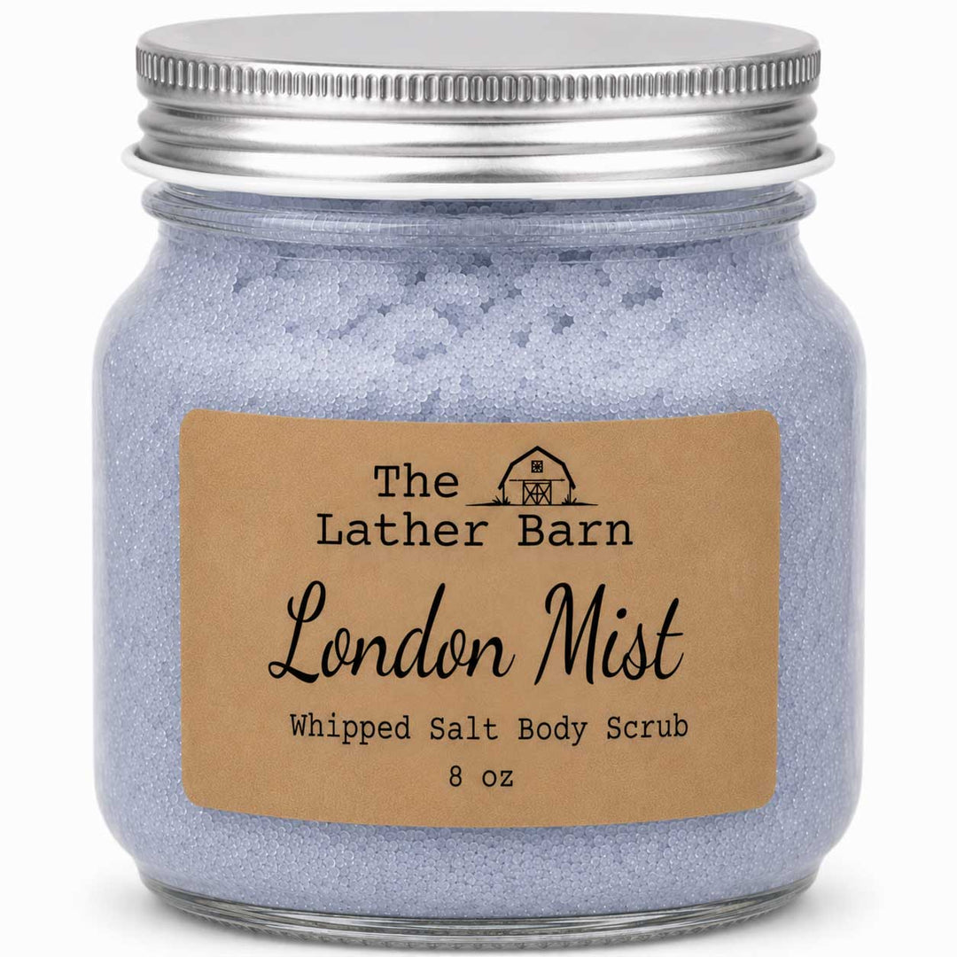 A jar of The Lather Barn London Mist Whipped Salt Body Scrub, featuring light lavender-blue scrub inside a clear glass jar with a silver metal lid and a brown kraft paper label.