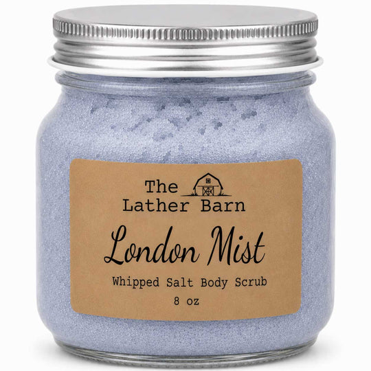 A jar of The Lather Barn London Mist Whipped Salt Body Scrub, featuring light lavender-blue scrub inside a clear glass jar with a silver metal lid and a brown kraft paper label.