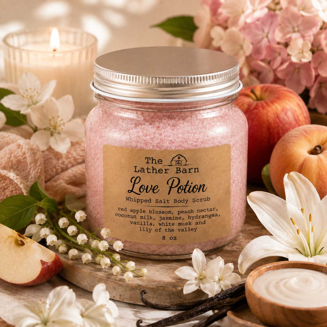 A jar of The Lather Barn Love Potion whipped salt body scrub sits on a wooden board surrounded by red apples, white lilies, flowers, a lit candle, and a small bowl of cream in a warm, styled setting.