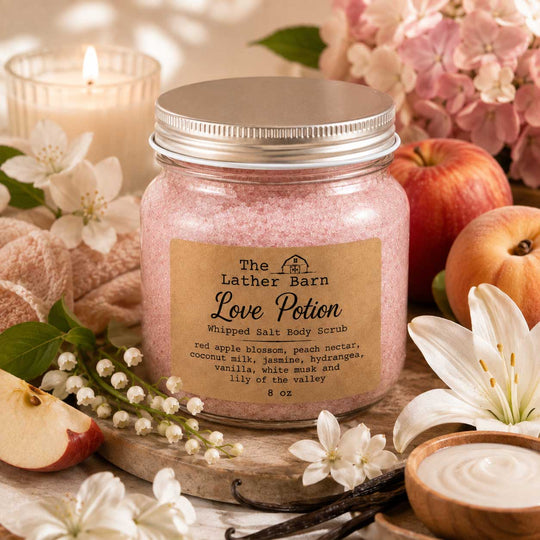 A jar of The Lather Barn Love Potion whipped salt body scrub sits on a wooden board surrounded by red apples, white lilies, flowers, a lit candle, and a small bowl of cream in a warm, styled setting.