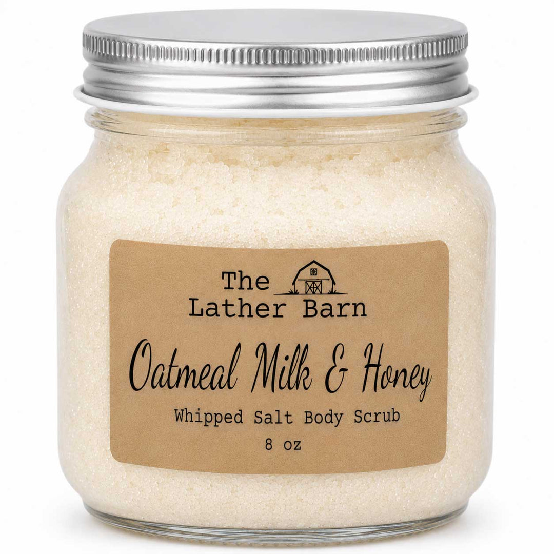 A glass jar of whipped salt body scrub with a silver metal lid and a brown kraft paper label reading 'The Lather Barn Oatmeal Milk & Honey Whipped Salt Body Scrub 8 oz'.