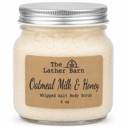 A glass jar of whipped salt body scrub with a silver metal lid and a brown kraft paper label reading 'The Lather Barn Oatmeal Milk & Honey Whipped Salt Body Scrub 8 oz'.