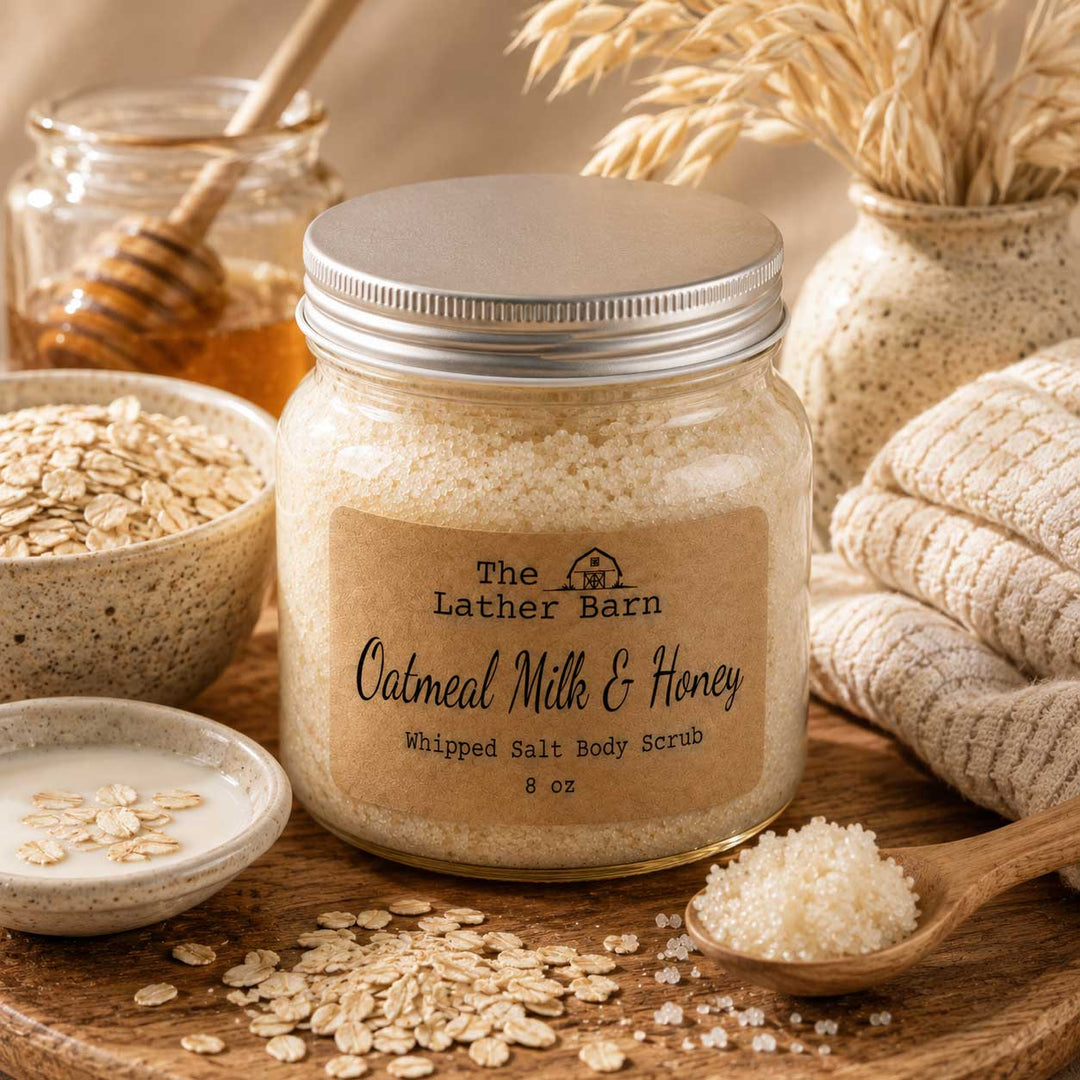 A glass jar of whipped salt body scrub by The Lather Barn sits on a wooden surface with oatmeal flakes, oats, honey, a folded cream towel, and dried wheat in the background, creating a warm, rustic spa-like setting.