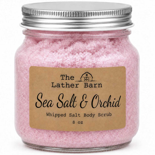 A clear glass jar filled with pink whipped salt body scrub, sealed with a silver metal lid, and labeled 'The Lather Barn Sea Salt & Orchid Whipped Salt Body Scrub 8 oz' on a white background.