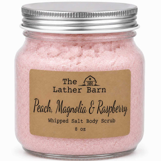 A glass jar filled with pink whipped salt body scrub, sealed with a silver metal lid and labeled 'The Lather Barn Peach, Magnolia & Raspberry Whipped Salt Body Scrub 8 oz' on a white background.