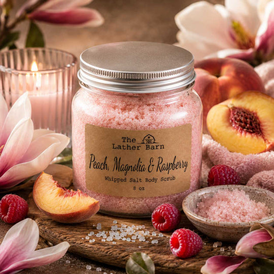 A glass jar of The Lather Barn Peach, Magnolia & Raspberry whipped salt body scrub sits on a rustic wooden board surrounded by fresh raspberries, sliced peaches, and soft pink flowers. A lit candle and a small bowl of pink salt crystals are visible in the background, creating a warm, inviting spa-like setting.