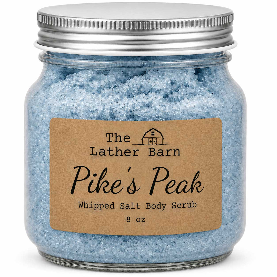 A glass jar filled with light blue whipped salt body scrub, sealed with a silver metal lid and featuring a brown kraft paper label that reads 'The Lather Barn Pike's Peak Whipped Salt Body Scrub 8 oz'.