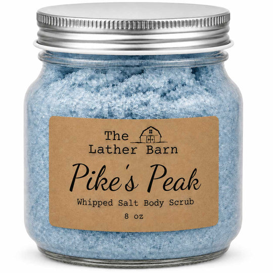 A glass jar filled with light blue whipped salt body scrub, sealed with a silver metal lid and featuring a brown kraft paper label that reads 'The Lather Barn Pike's Peak Whipped Salt Body Scrub 8 oz'.
