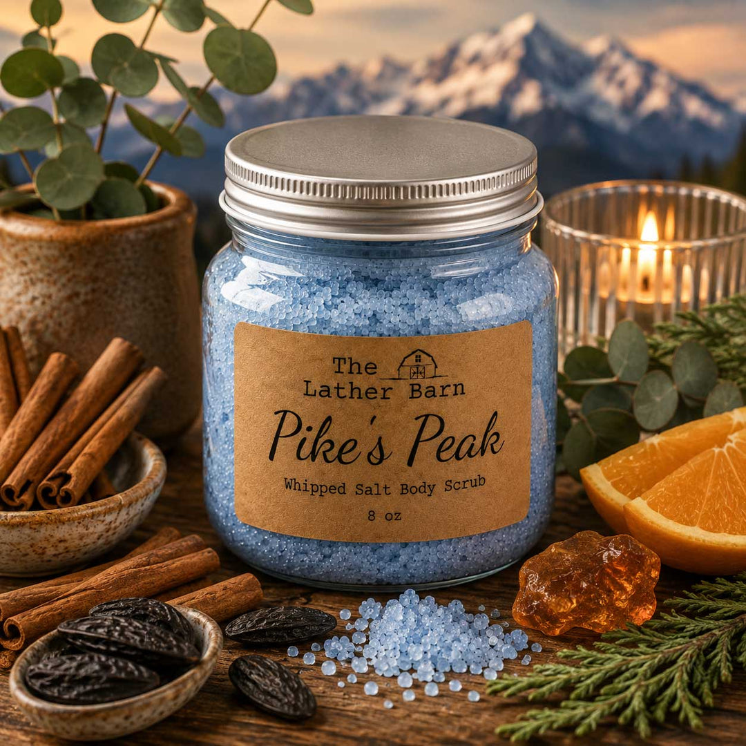 A jar of whipped salt body scrub by The Lather Barn, labeled 'Pike's Peak', filled with light blue salt crystals and topped with a silver lid. The jar is styled on a rustic wooden surface with cinnamon sticks, eucalyptus, a lit candle, and orange slices, with a mountain landscape in the background.