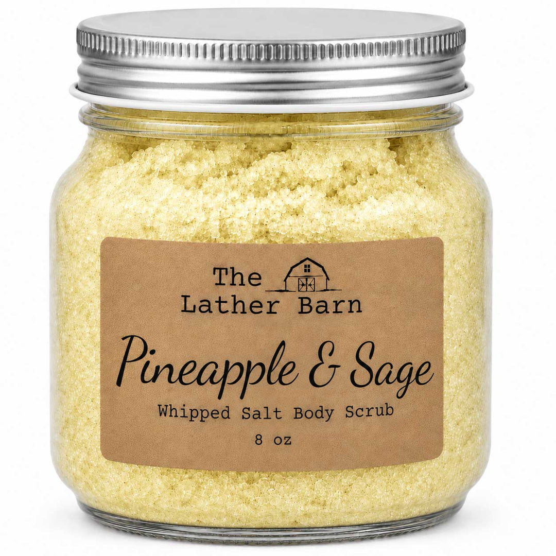 A glass jar of The Lather Barn Pineapple & Sage Whipped Salt Body Scrub, filled with pale yellow scrub and sealed with a silver metal lid, shown on a white background.