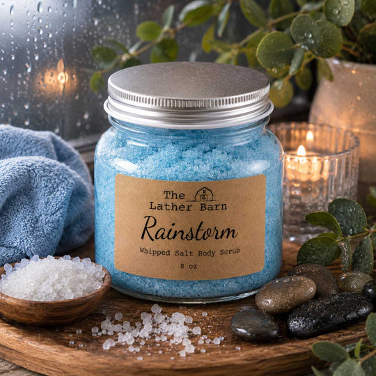 A glass jar filled with light blue whipped salt body scrub, sealed with a silver metal lid and featuring a brown kraft paper label that reads 'The Lather Barn Rainstorm Whipped Salt Body Scrub 8 oz'.