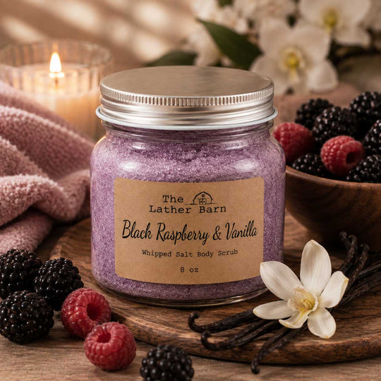 A jar of The Lather Barn Black Raspberry & Vanilla Whipped Salt Body Scrub sits on a wooden board surrounded by fresh raspberries, blackberries, vanilla beans, and a white flower, with a soft pink towel and a lit candle blurred in the background.