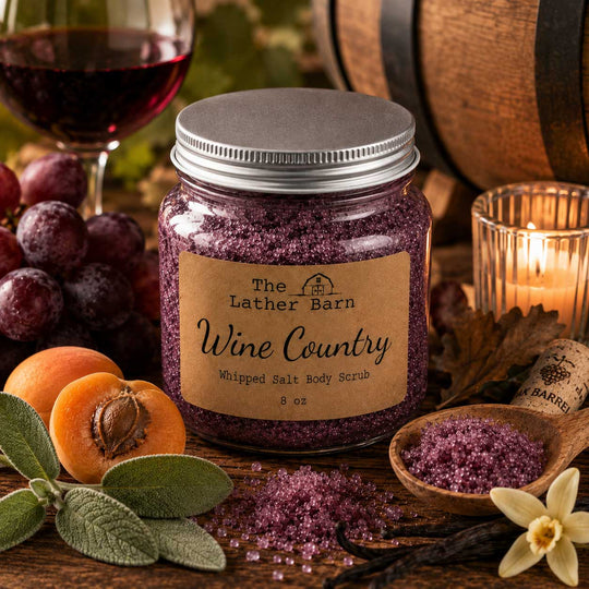 A jar of The Lather Barn Wine Country Whipped Salt Body Scrub sits on a rustic wooden table surrounded by wine glass, grapes, apricots, sage leaves, vanilla beans, and a lit candle, with loose purple scrub visible in the foreground.