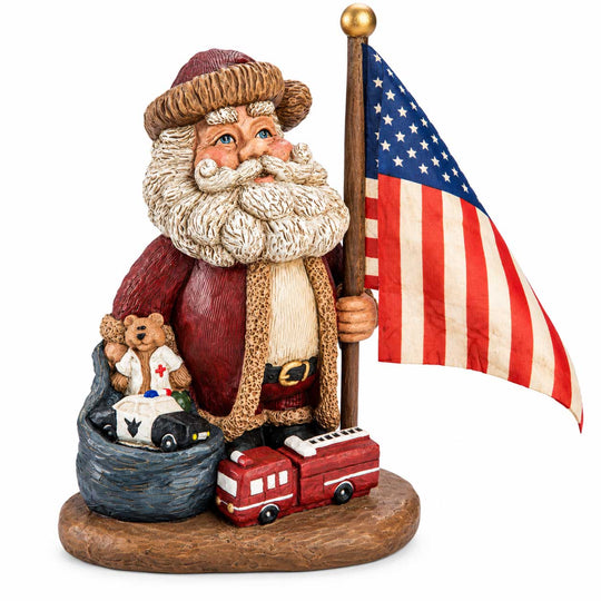 A Santa Claus figurine wearing a firefighter outfit, holding a US flag on a gold pole. Santa is standing on a brown base next to a red fire truck and a teddy bear in a bag wearing a white firefighter jacket with a cross.