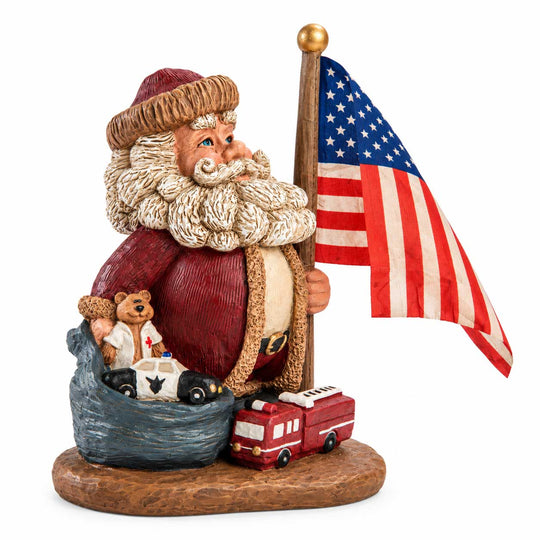 A Santa Claus figurine holding an American flag, with a red Santa suit, white beard, brown hat, and a base featuring a small teddy bear in a police car and a red fire truck, photographed on a white background.