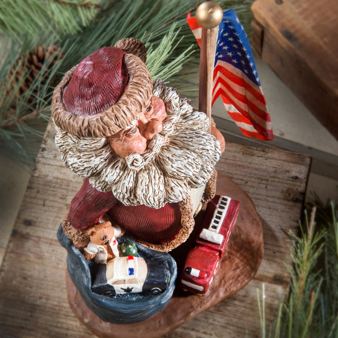Decorative figure of Santa with a hat, holding an American flag, with a small boat and toy truck below.