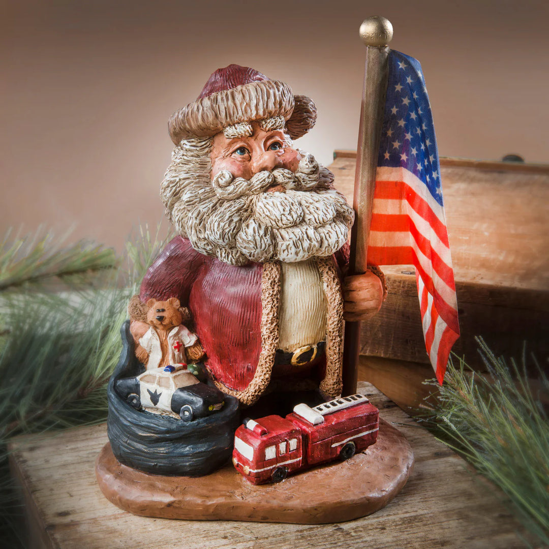 Decorative figurine of Santa with a flag, surrounded by small vehicles and a teddy bear on a wooden base.