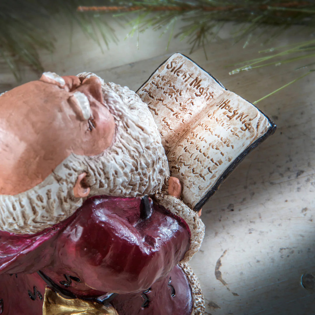 Statue of Santa in a red garment with a book titled 'Silent Night' against a natural background.