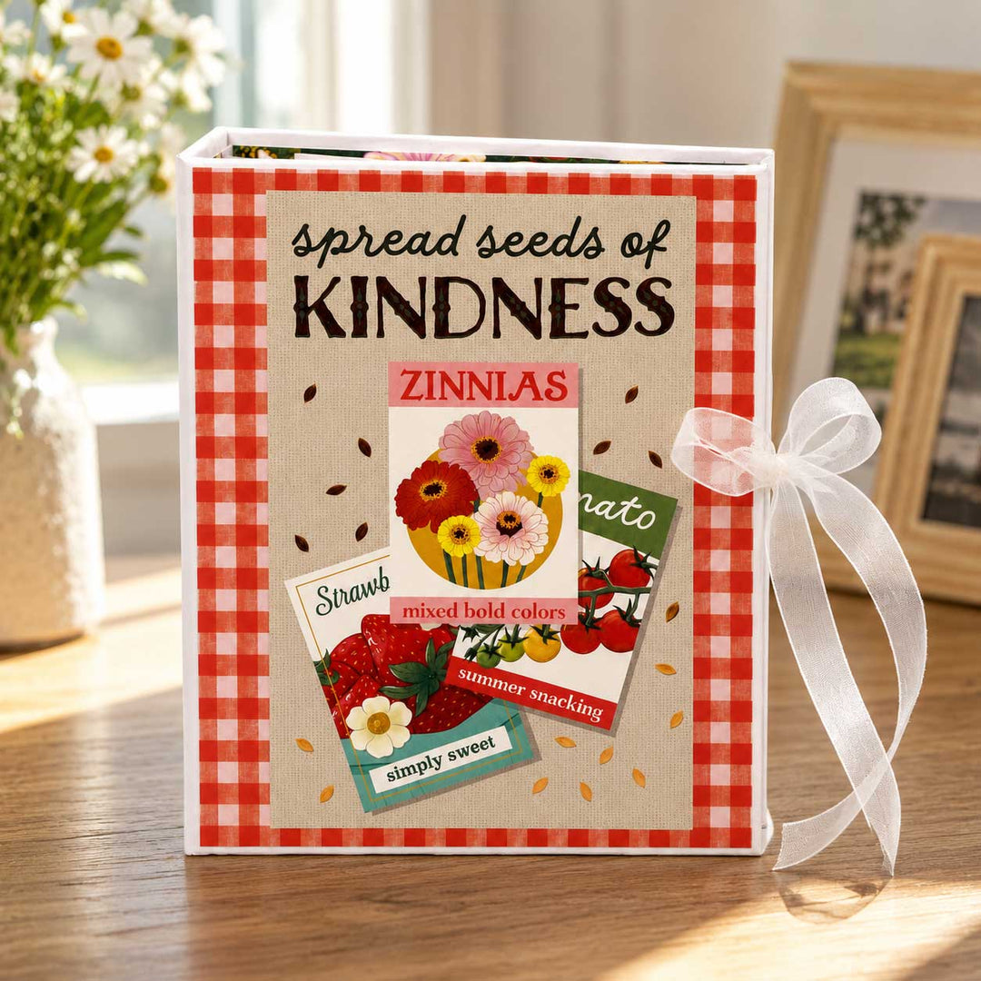 A decorative seed starter journal or book titled 'spread seeds of KINDNESS' with a red gingham border, colorful zinnia flower illustration, and a white ribbon bow on the side, placed on a wooden table in a softly lit indoor setting.