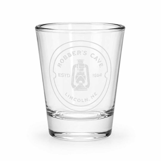 A clear glass shot glass featuring a frosted white circular stamp that reads 'Robber's Cave Estd 1864 Lincoln, NE' with a lantern graphic, set against a plain white background.