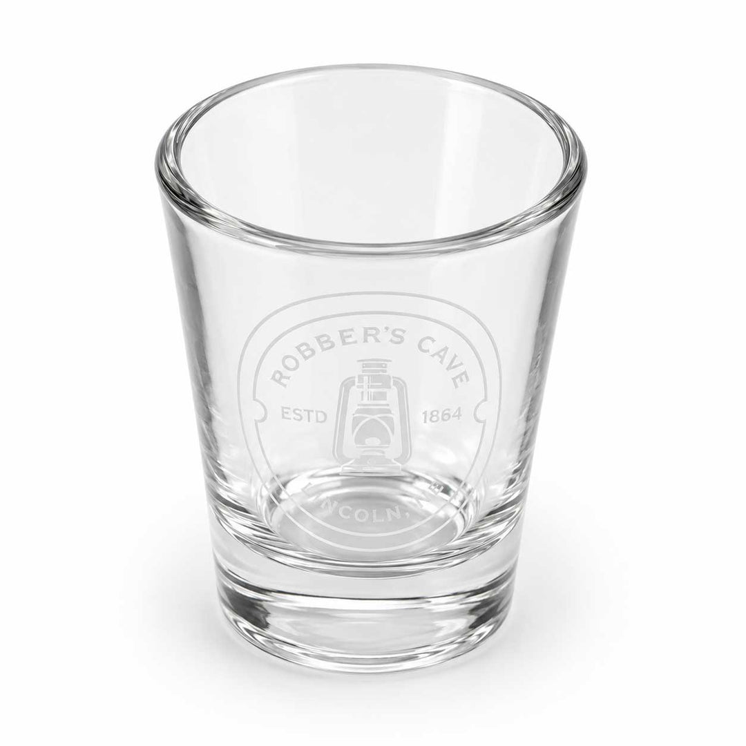 A clear glass shot glass featuring an etched circular logo that reads 'ROBBER'S CAVE ESTD 1664' with a lantern illustration, set against a plain white background.