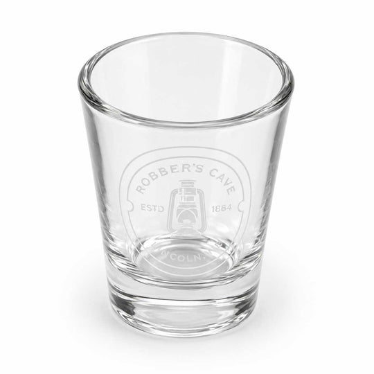 A clear glass shot glass featuring an etched circular logo that reads 'ROBBER'S CAVE ESTD 1664' with a lantern illustration, set against a plain white background.
