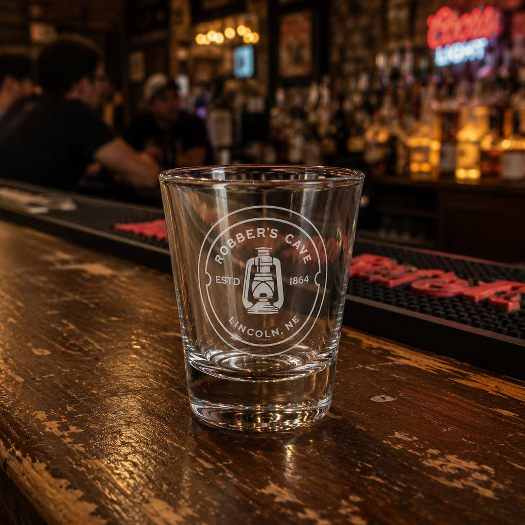 A clear glass tumbler with a frosted white circular logo reading 'Robber's Cafe Lincoln, NE' and a lantern graphic, set on a rustic wooden bar counter in a blurred bar interior.
