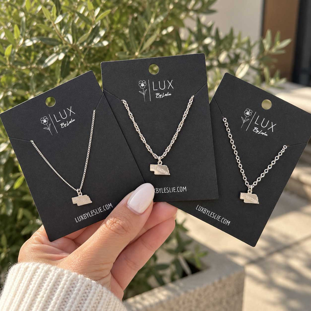 Three silver necklace cards with small state-shaped pendants are held in a hand outdoors in front of greenery. The cards are branded LUX by Leslie.