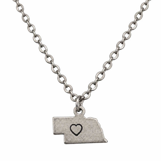 A silver-toned chain necklace with a pendant shaped like the state of Nebraska, featuring a small black heart outline in the center, shown on a plain white background.