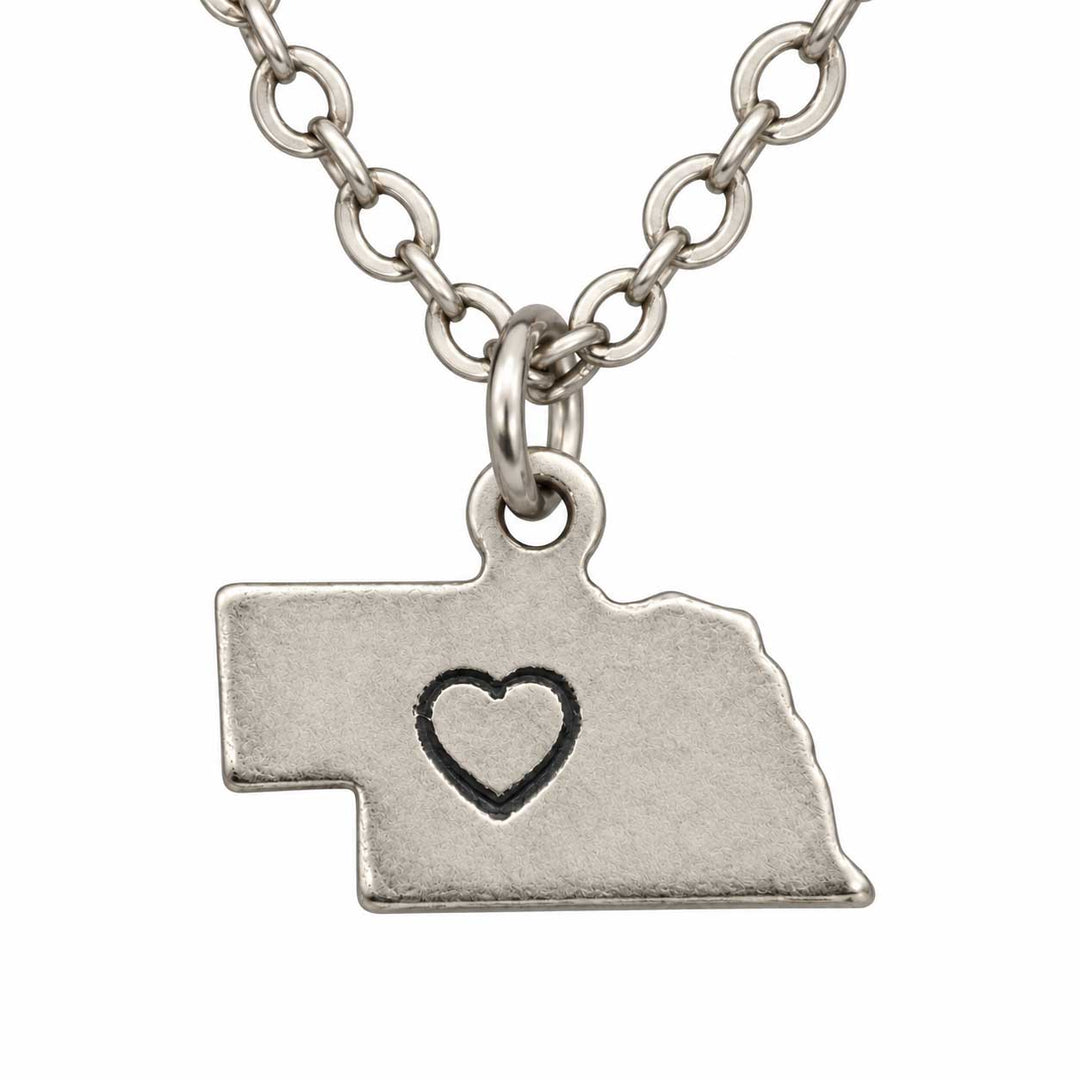 A silver-toned chain necklace with a pendant shaped like the state of Nebraska, featuring a small black heart outline in the center, shown on a plain white background.