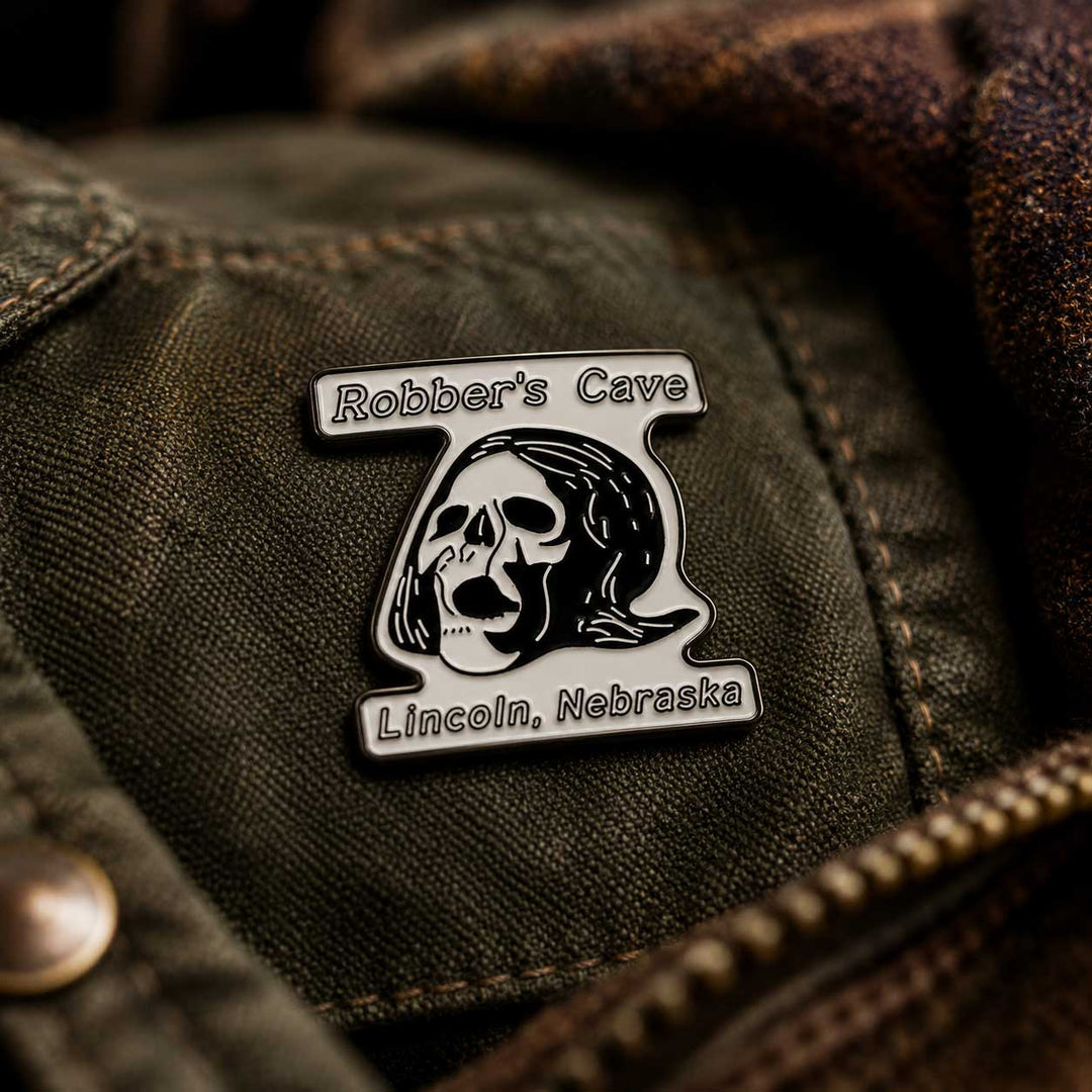 A close-up of a metal enamel pin featuring a skull and the text 'Robber's Cave' and 'Lincoln, Nebraska', attached to an olive green denim jacket.
