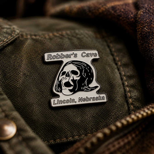 A close-up of a metal enamel pin featuring a skull and the text 'Robber's Cave' and 'Lincoln, Nebraska', attached to an olive green denim jacket.