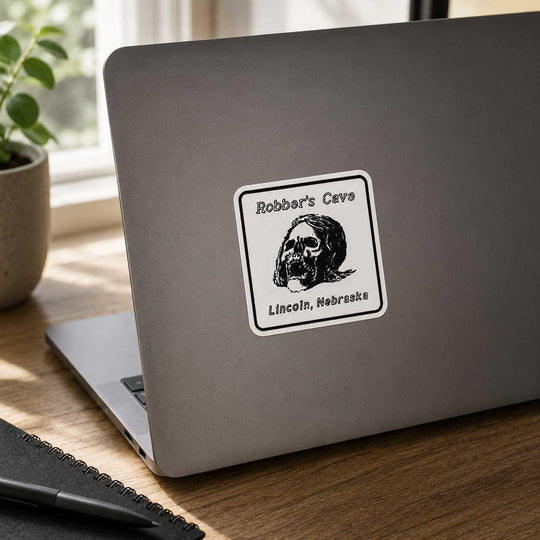 A square white sticker with a black border and illustration of a cave skull, reading 'Robber's Cave' and 'Lincoln, Nebraska,' applied to the back of a closed silver laptop on a wooden desk near a window.
