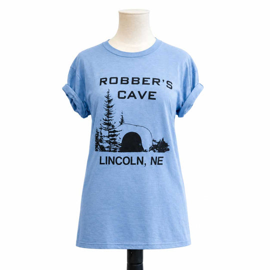 A light blue heather t-shirt displayed on a mannequin torso against a white background. The shirt features a black graphic print with the text 'ROBBER'S CAVE' and 'LINCOLN, NE' surrounding a silhouette of a campfire and trees.