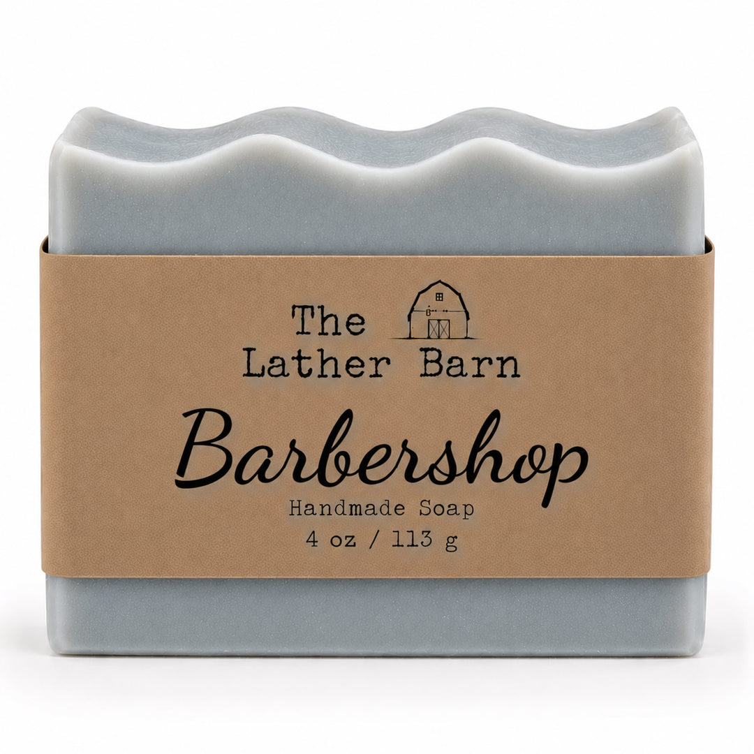 A rectangular bar of handmade soap in a pale blue-gray color, wrapped in a brown kraft paper band with black text that reads 'The Lather Barn Barbershop Handmade Soap 4 oz / 113 g'.