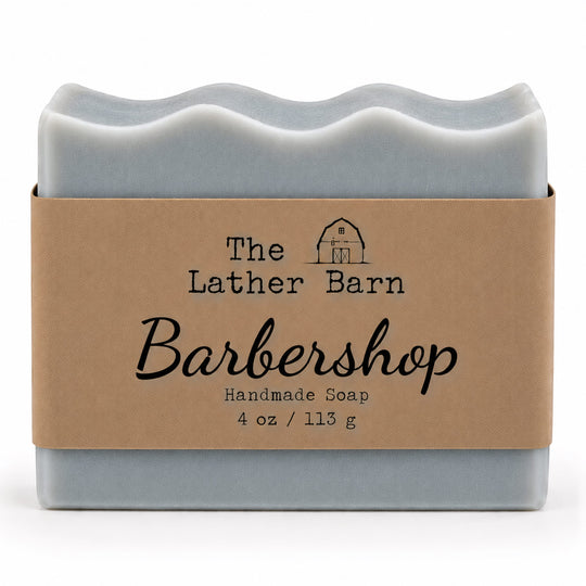 A rectangular bar of handmade soap in a pale blue-gray color, wrapped in a brown kraft paper band with black text that reads 'The Lather Barn Barbershop Handmade Soap 4 oz / 113 g'.