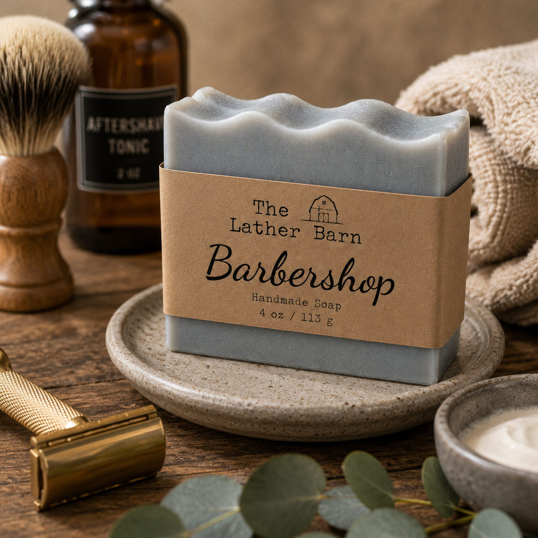 A rectangular bar of handmade soap in a dusty blue-gray color sits on a ceramic dish, wrapped in a kraft paper band reading 'The Lather Barn Barbershop.' The scene is styled with barbershop-themed props including a brass safety razor, shaving brush, amber bottle, and folded towel on a wooden surface.