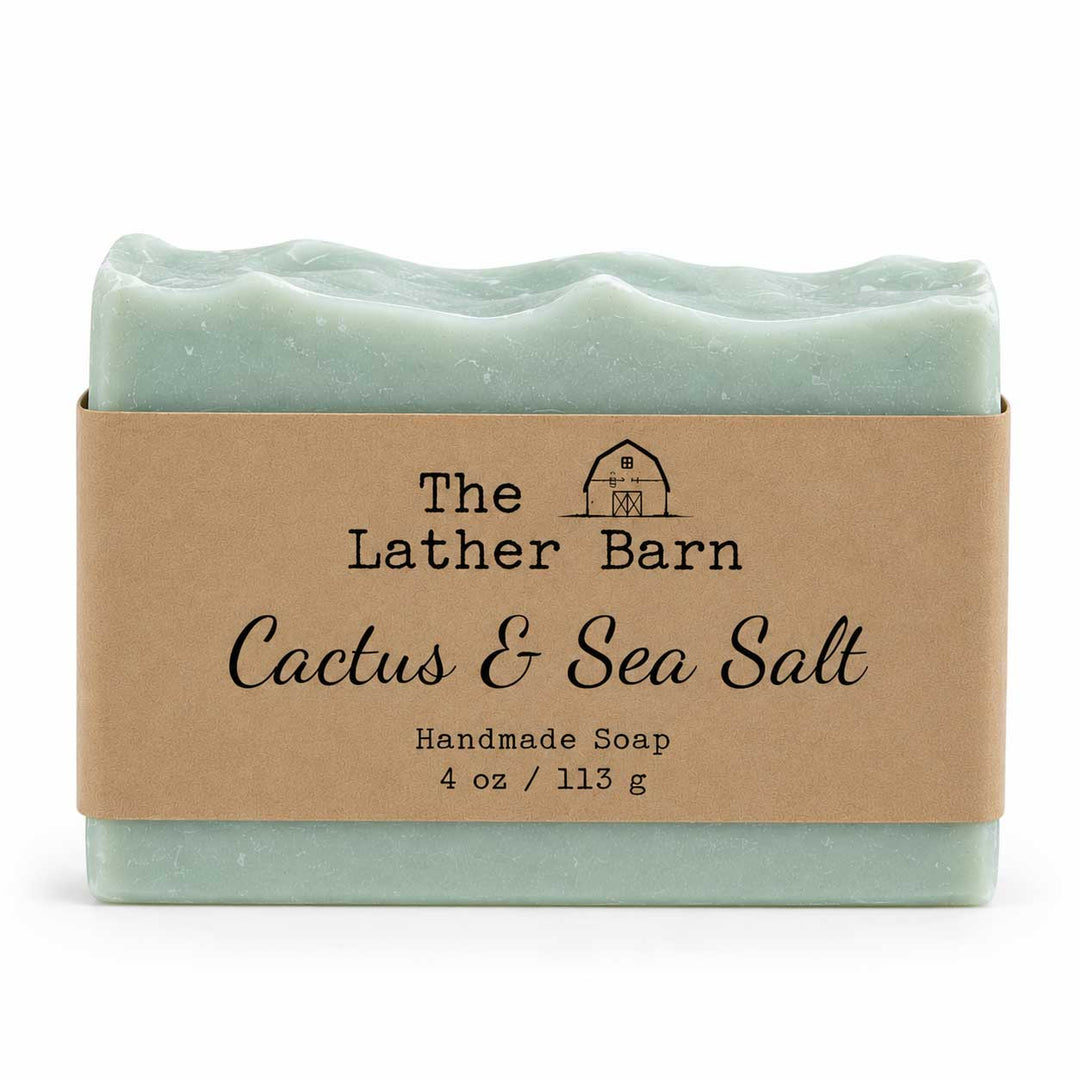 A handmade soap bar wrapped in a kraft paper band. The soap is a pale mint green with a wavy edge, and the label reads 'The Lather Barn Cactus & Sea Salt Handmade Soap 4 oz / 113 g'.