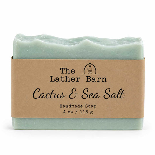 A handmade soap bar wrapped in a kraft paper band. The soap is a pale mint green with a wavy edge, and the label reads 'The Lather Barn Cactus & Sea Salt Handmade Soap 4 oz / 113 g'.
