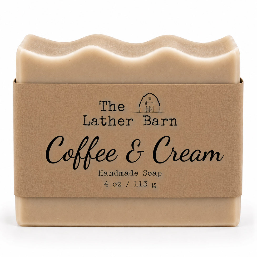 A 4 oz bar of handmade soap in a kraft paper sleeve. The soap is a light beige color with a wavy top edge, and the sleeve features the brand name 'The Lather Barn' and the scent name 'Coffee & Cream'.