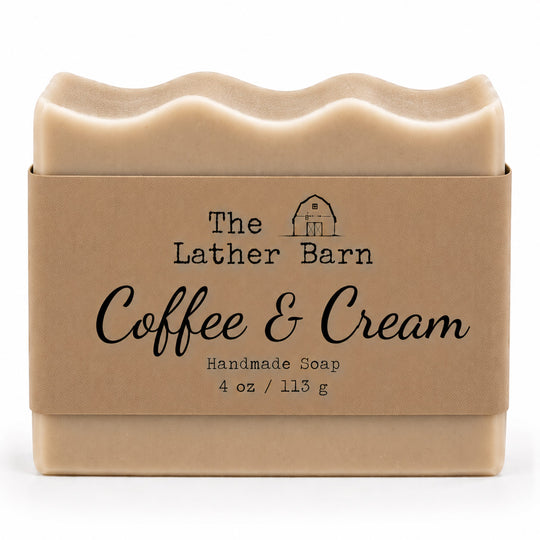 A 4 oz bar of handmade soap in a kraft paper sleeve. The soap is a light beige color with a wavy top edge, and the sleeve features the brand name 'The Lather Barn' and the scent name 'Coffee & Cream'.