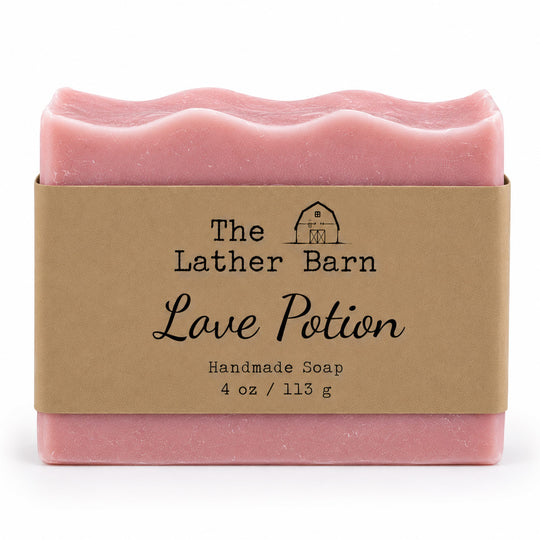 A pink handmade soap bar wrapped in a kraft paper band labeled 'The Lather Barn Love Potion Handmade Soap 4 oz / 113 g' on a white background.