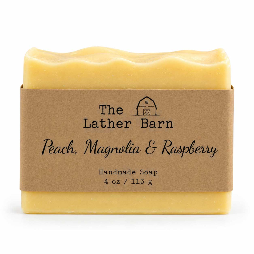 A rectangular bar of handmade soap in pale peach-yellow with a wavy top edge, wrapped in a brown kraft paper band labeled 'The Lather Barn Peach, Magnolia & Raspberry Handmade Soap 4 oz / 113 g' on a white background.