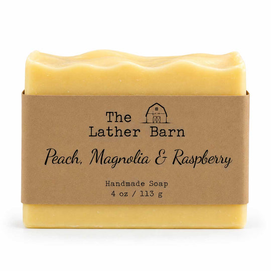 A rectangular bar of handmade soap in pale peach-yellow with a wavy top edge, wrapped in a brown kraft paper band labeled 'The Lather Barn Peach, Magnolia & Raspberry Handmade Soap 4 oz / 113 g' on a white background.