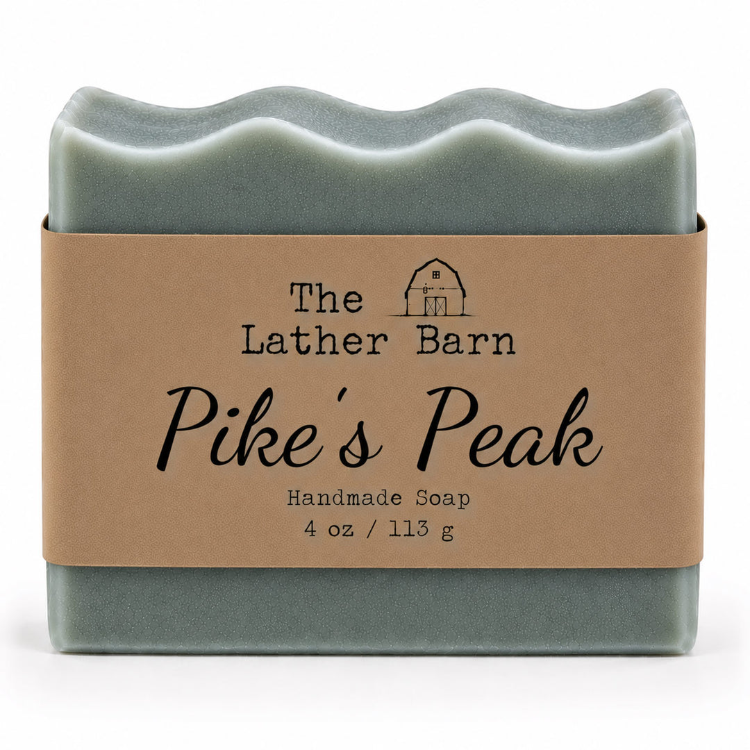 A rectangular handmade soap bar wrapped in a kraft paper band. The soap is a muted sage green, and the label reads 'The Lather Barn Pike's Peak Handmade Soap 4 oz / 113 g' with a small barn illustration.