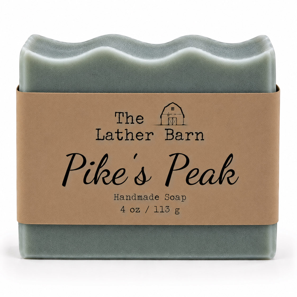 A rectangular handmade soap bar wrapped in a kraft paper band. The soap is a muted sage green, and the label reads 'The Lather Barn Pike's Peak Handmade Soap 4 oz / 113 g' with a small barn illustration.
