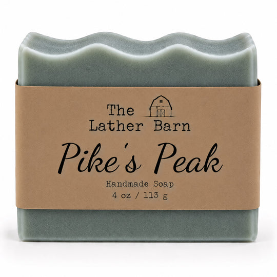 A rectangular handmade soap bar wrapped in a kraft paper band. The soap is a muted sage green, and the label reads 'The Lather Barn Pike's Peak Handmade Soap 4 oz / 113 g' with a small barn illustration.