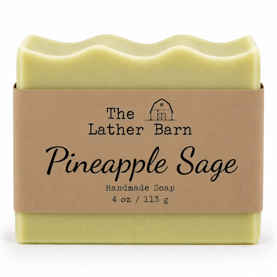 A 4 oz bar of handmade soap by The Lather Barn, named Pineapple Sage. The soap is pale yellow-green and wrapped in a brown kraft paper band with black text and a small barn logo, set against a plain white background.